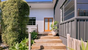 Una casa moderna con terraza de madera que conduce a una puerta principal, rodeada de follaje y una valla de madera.