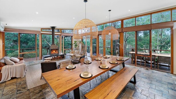 A spacious dining area with a large wooden table set for a meal, surrounded by large windows offering a view of the forest. - Kurraburra Lodge (Anglesea)