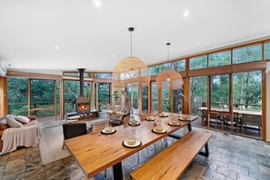 Une vaste salle à manger avec une grande table en bois dressée pour le repas, entourée de grandes fenêtres offrant une vue sur la forêt.
