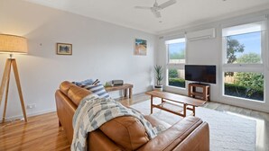 A spacious living room with a brown leather sofa, a wooden coffee table, a floor lamp, a television, and large windows offering a view of greenery. - Secluded Anderson - in the heart of Torquay (Torquay)