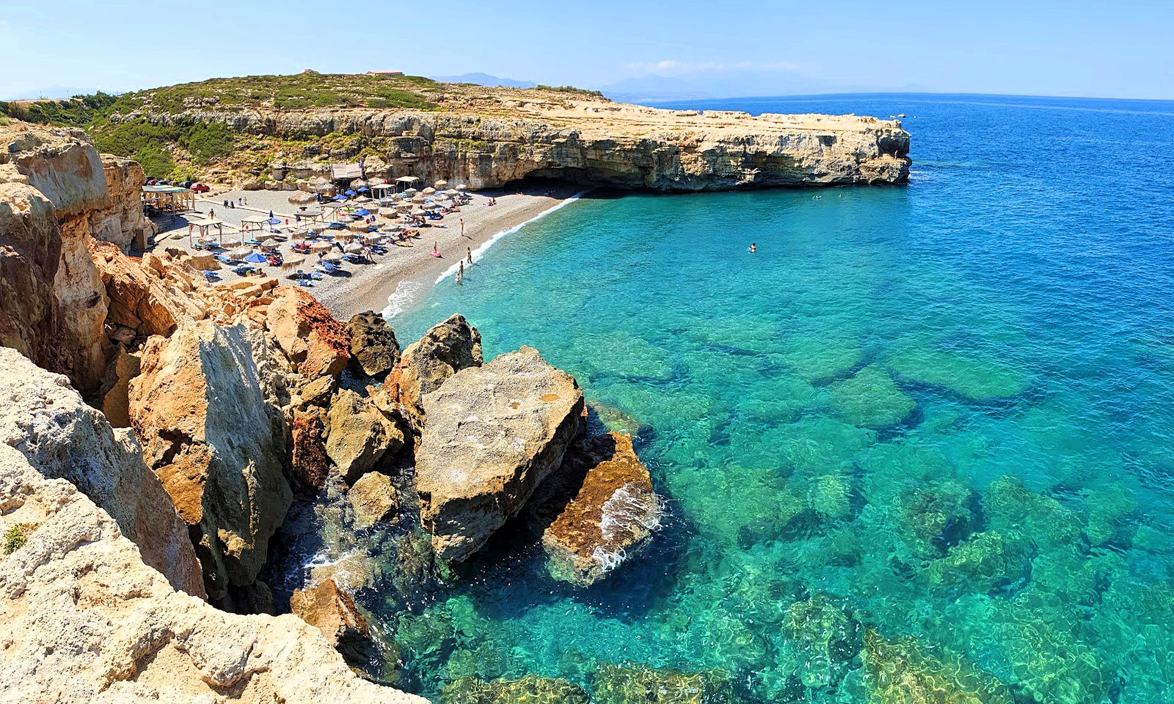 A coastal beach with clear turquoise waters, rocky cliffs, and a sandy beach area with umbrellas and sun loungers.