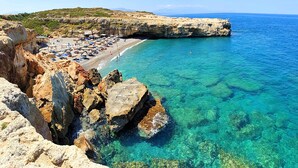 Una playa costera con aguas turquesas cristalinas, acantilados rocosos y una zona de arena con sombrillas y tumbonas.