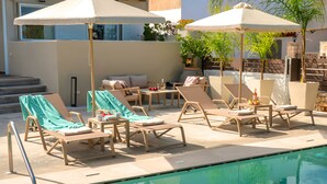 Zona junto a la piscina con sillones reclinables y sombrillas, una pequeña mesa con bebidas, y vista a la piscina.