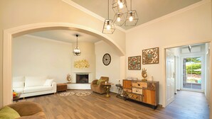 Un salón amplio con un sofá blanco, un sillón marrón y un aparador de madera. Hay una chimenea, un candelabro y obras de arte en las paredes.