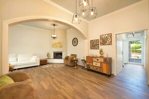A spacious living room with a white sofa, a brown armchair, and a wooden cabinet. There is a fireplace, a chandelier, and artwork on the walls. - Villa di Capaka Private Pool (Roupes)