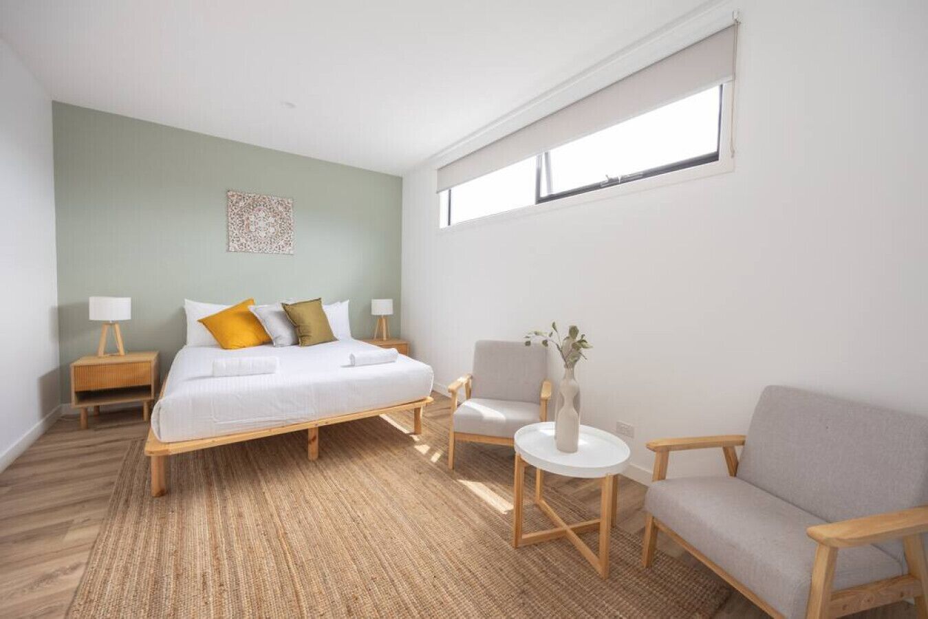 Ein modernes Hotelzimmer mit einem großen Bett, zwei Nachttischen, einem kleinen runden Tisch mit einer Vase und einem grauen Sofa.