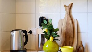 A kitchen with white cabinets, a stainless steel kettle, a green vase with lemons, a wooden cutting board, and a yellow bowl. - Cosy living at Cosy corner (Torquay)