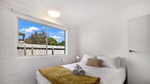 A hotel room with a large window, white brick walls, a bed with white bedding and a mustard-colored throw blanket, and a small bedside table with a lamp. - Cosy living at Cosy corner (Torquay)