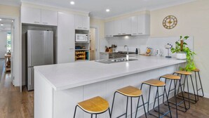 A modern kitchen with white cabinetry, a stainless steel refrigerator, a microwave, and a sink. There are four wooden stools at the island. - Barwon Bliss - Ocean Grove (Ocean Grove)