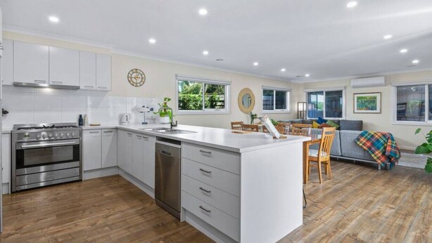 A modern kitchen with white cabinets, a stainless steel stove, and a dishwasher. There is a dining area with a wooden table and chairs, and a sofa with a colorful blanket. - Barwon Bliss - Ocean Grove (Ocean Grove)