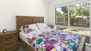A bedroom with a wooden bed, a nightstand, and a large window with a view of a wooden fence and trees. - Beach Breeze Bungalow - in the heart of Torquay (Torquay)