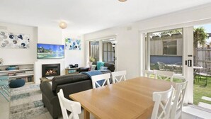 A modern living room with a fireplace, a dining table, and a large window overlooking a garden. - Beach Breeze Bungalow - in the heart of Torquay (Torquay)