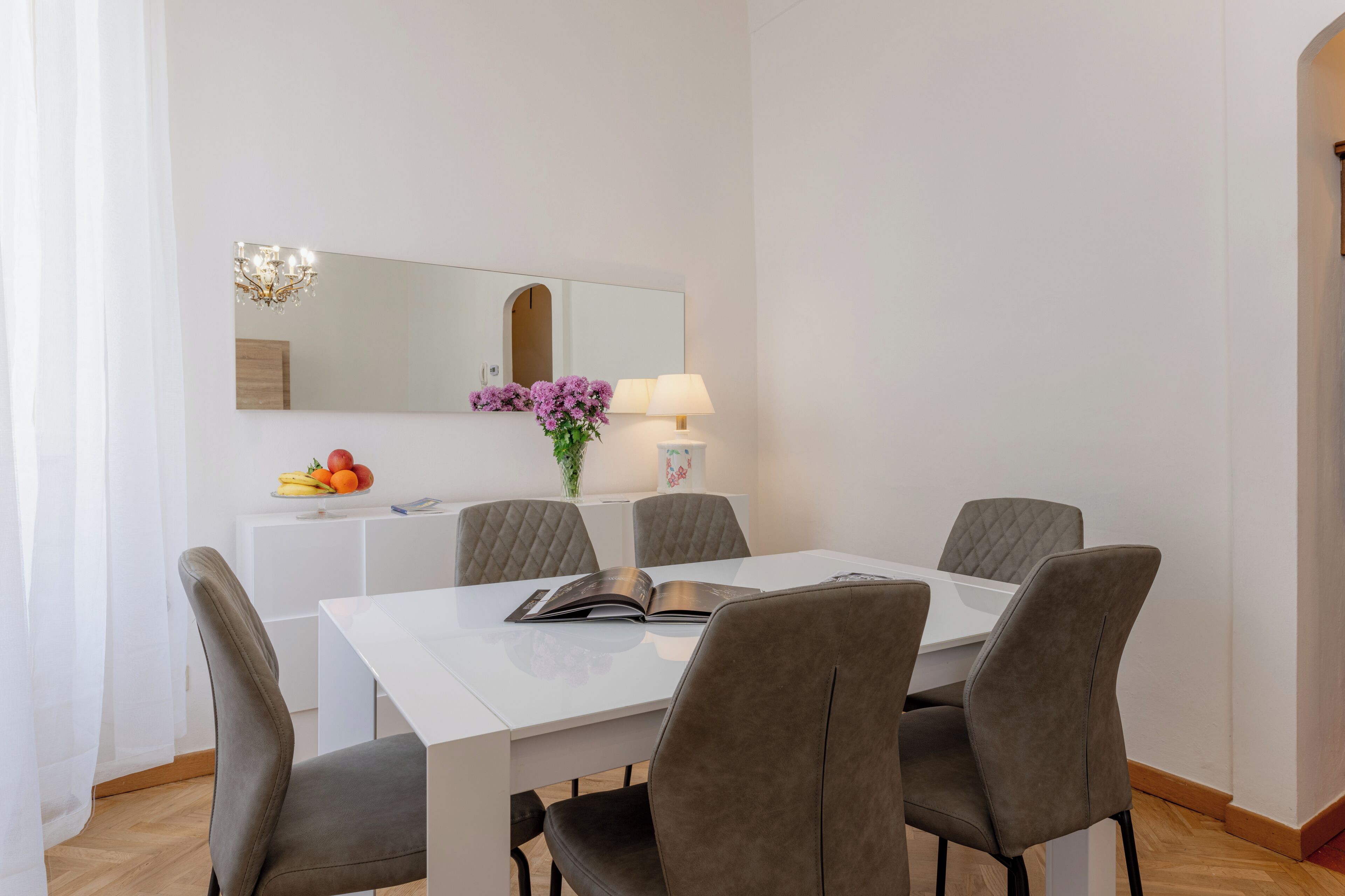 Uma sala de jantar moderna com uma mesa branca, quatro cadeiras cinzas estofadas, um armário branco com um espelho acima, um vaso com flores roxas e uma fruteira no armário.
