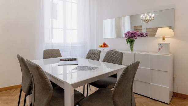 Un comedor moderno con una mesa blanca, sillas tapizadas en gris, un armario blanco con espejo y un jarrón con flores.
