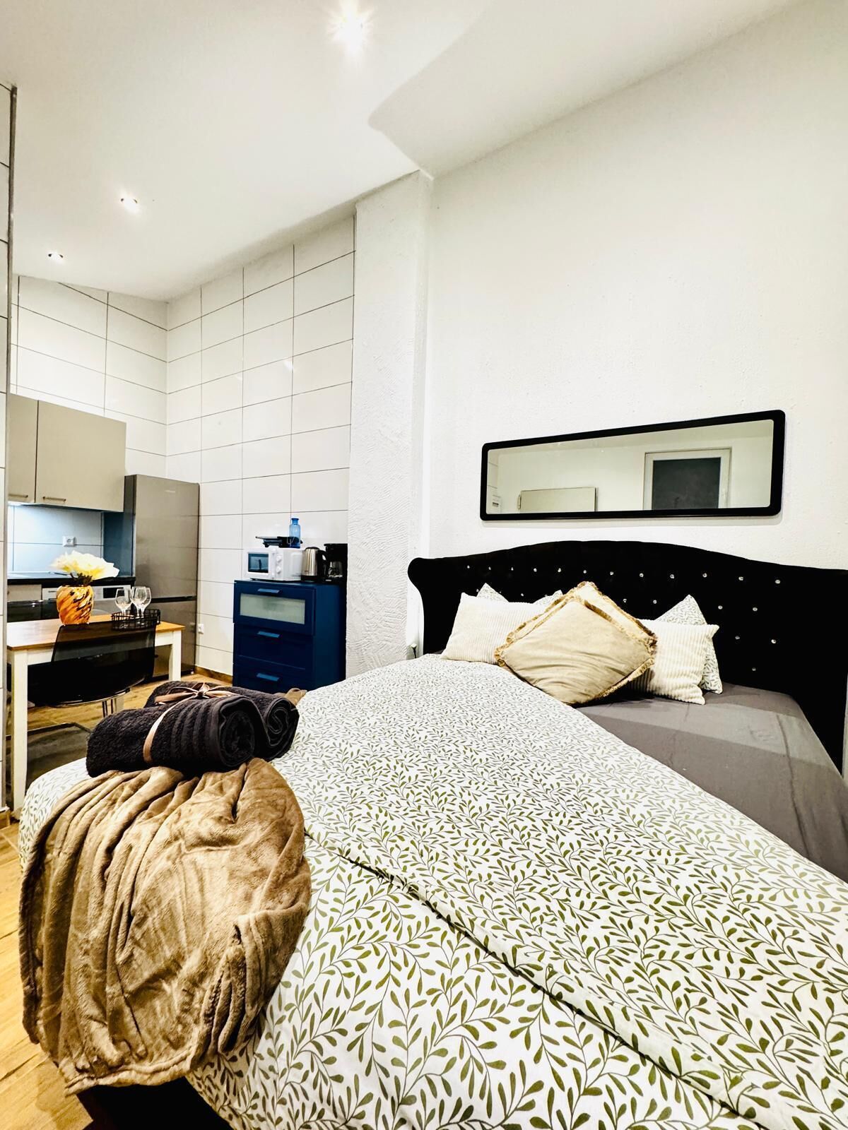 Una habitación de hotel moderna con una cama bien hecha, una kitchenette pequeña y un espejo grande en la pared.