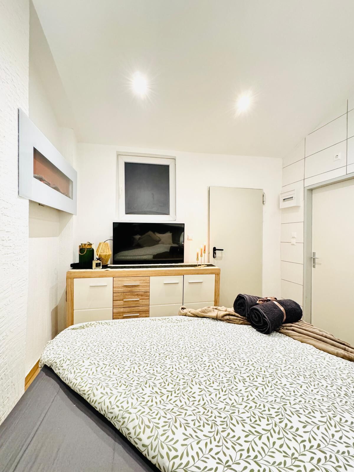 A bedroom with a bed, a TV on a wooden dresser, a framed picture, and a mirror.