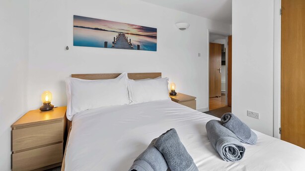 Una cama bien hecha con sábanas blancas y dos toallas enrolladas, una mesita de noche con una lámpara, un cuadro de un atardecer sobre un muelle y una puerta de madera.