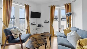 Una sala de estar moderna con un sofá azul, una mesa de centro de madera y un sillón azul. Hay un televisor de pantalla plana montado en la pared y una ventana con cortinas.