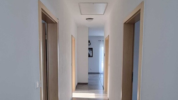 Un couloir étroit, doté d’un plancher en bois et de murs aux couleurs claires, qui mène à une pièce dont la porte se trouve au fond.