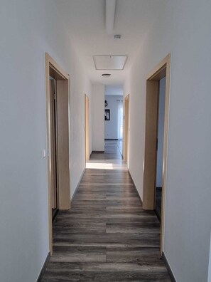A narrow hallway with wooden flooring and light-colored walls, leading to a room with a door at the end.