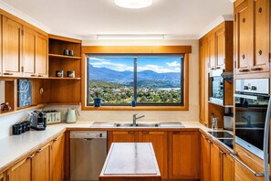 A modern kitchen with wooden cabinets, a large window with a scenic view, a built-in dishwasher, and a stainless steel oven. - The Lookout | Panoramic Views | Minutes to Hobart (Bellerive)
