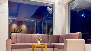 A modern living room with a beige sectional sofa, a wooden side table with a vase of flowers, and a large window with a view of the city. - HOTEL RIVA INN (Miraj)