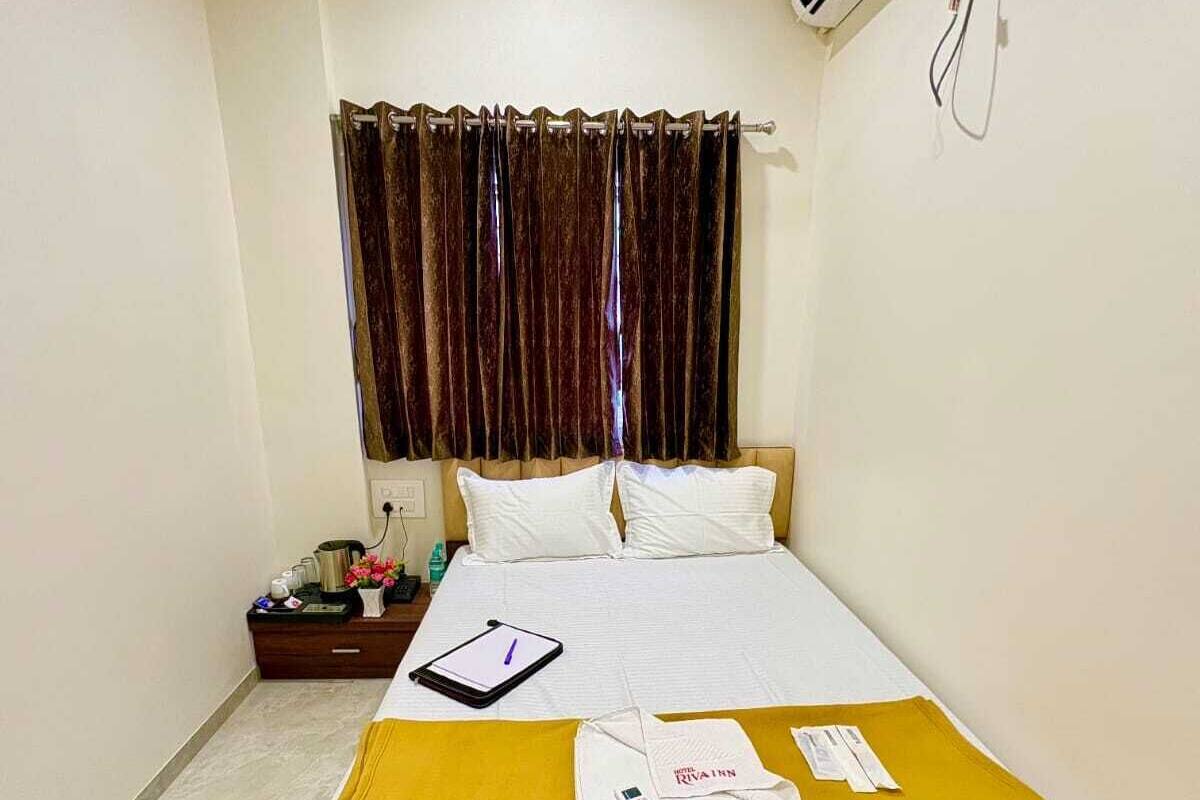 A hotel room with a bed, a bedside table, a nightstand, a window with curtains, and an air conditioning unit.