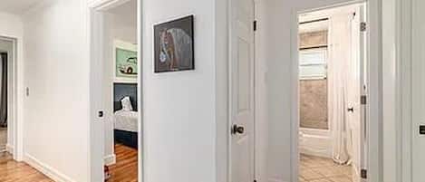 Un pasillo con piso de madera y paredes blancas que conduce a un dormitorio con cama y puerta.