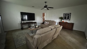A living room with a beige sofa, a flat-screen TV, a ceiling fan, and a small table with books and candles.
