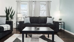 Ett vardagsrum med en mörkgrå soffa, två matchande kuddar, ett soffbord, en mönstrad matta, en krukväxt, ett sidobord med en lampa och ett fönster med vita gardiner.