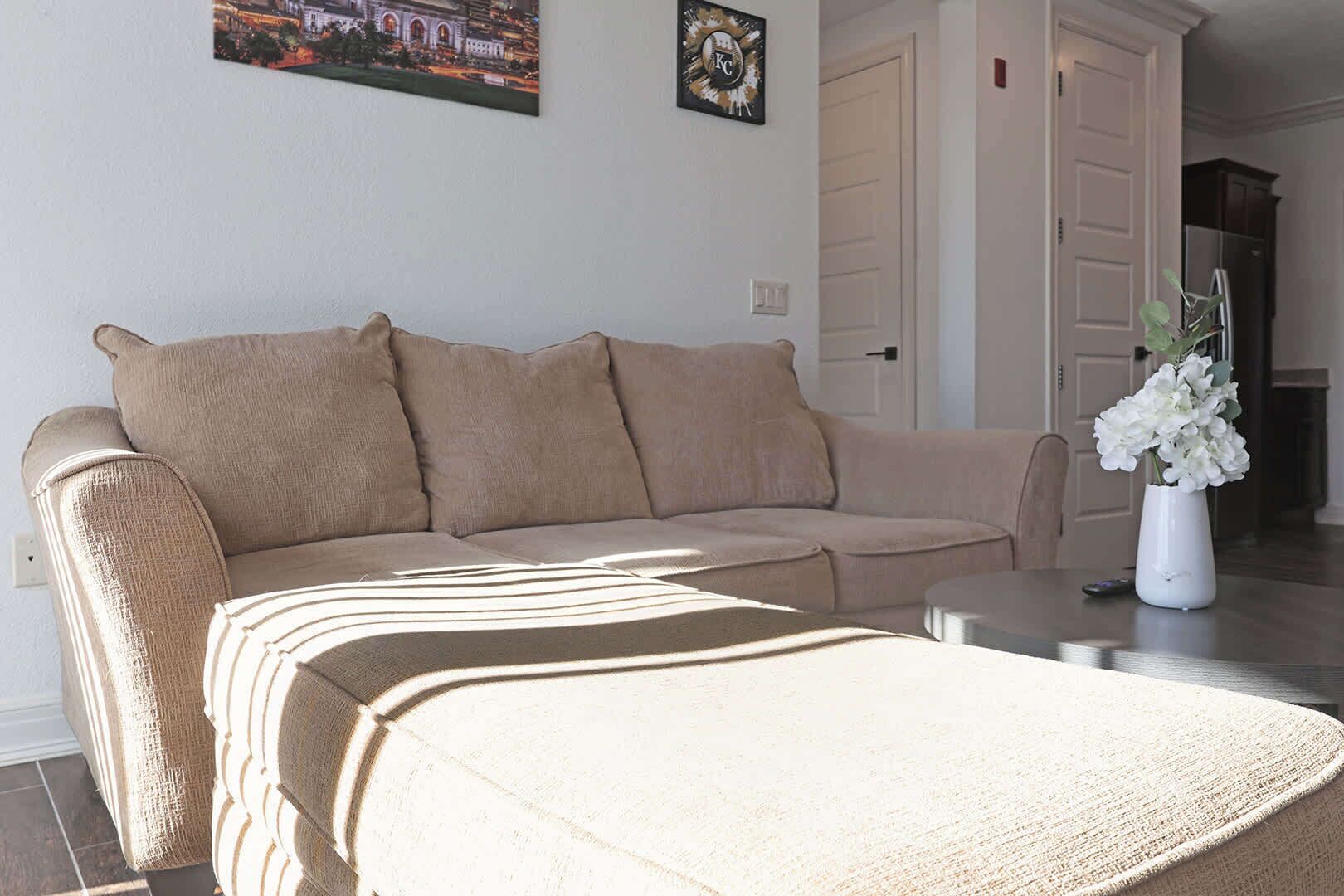 En beige soffa med ett randigt pläd, en vit vas med blommor och en målning av en stadsbild på väggen.
