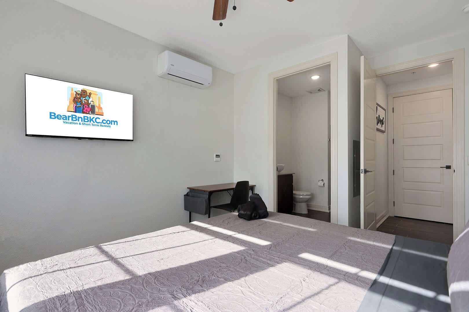 Une chambre à coucher comprenant un lit, un bureau, une chaise, un climatiseur et une salle de bain visible à travers une porte ouverte.