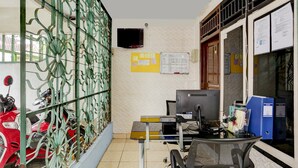A room with a desk, computer, and a chair. There is a window with a metal grill and a red scooter parked outside. - OYO 94908 Demang Kostel (Palembang)