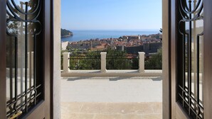 Porta aperta con cancelli in ferro battuto che conducono a un balcone con vista su una cittadina costiera con edifici storici e un cielo azzurro limpido.