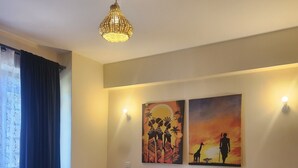 Un letto rifatto con una coperta bianca e arancione, una struttura in metallo nero e un cuscino decorativo. Sono presenti due applique e due quadri raffiguranti scene africane.