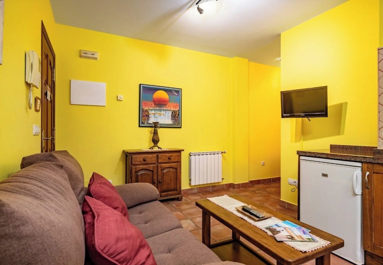 A living room with a sofa, a coffee table, a TV, a small kitchen area, and a painting on the wall.