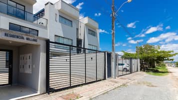 Un edificio residencial moderno con entrada protegida por un portón, un cuadro eléctrico visible y cielo despejado.
