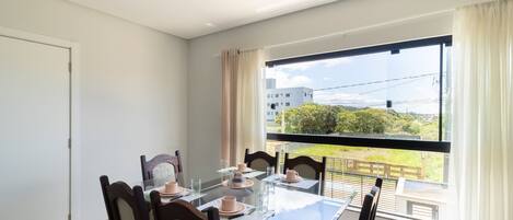 A dining area with a glass table and chairs, a view of greenery and buildings through a window.