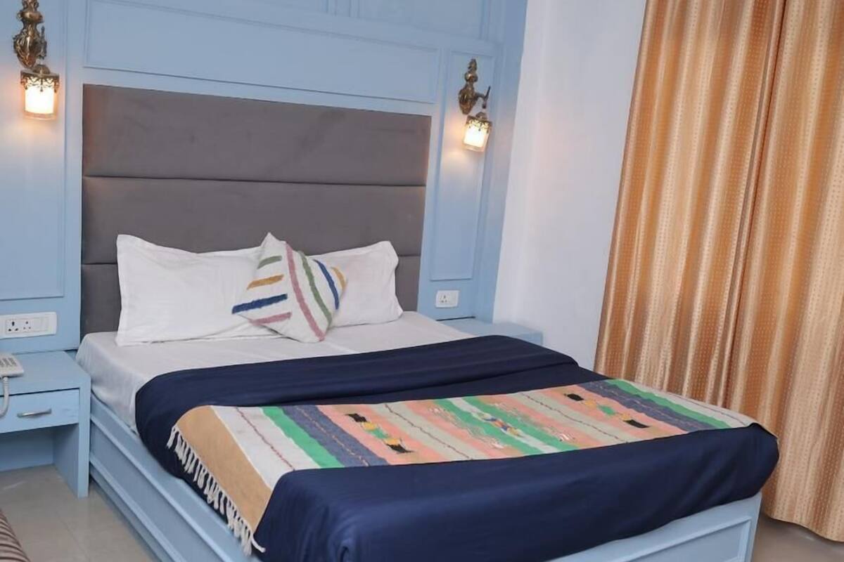 A bedroom with a light blue headboard, a bed with a colorful blanket, a striped bench, and a curtain.