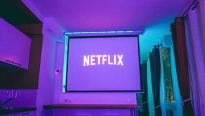 Una habitación con una cama, un taburete pequeño, una pantalla en la pared que muestra “NETFLIX” y una cesta colgante del techo.