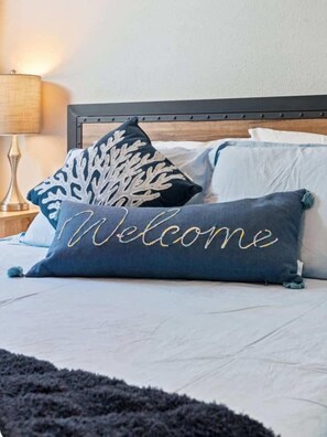 A bed with a 'Welcome' pillow, a decorative pillow with a leaf pattern, and a lamp on a bedside table.