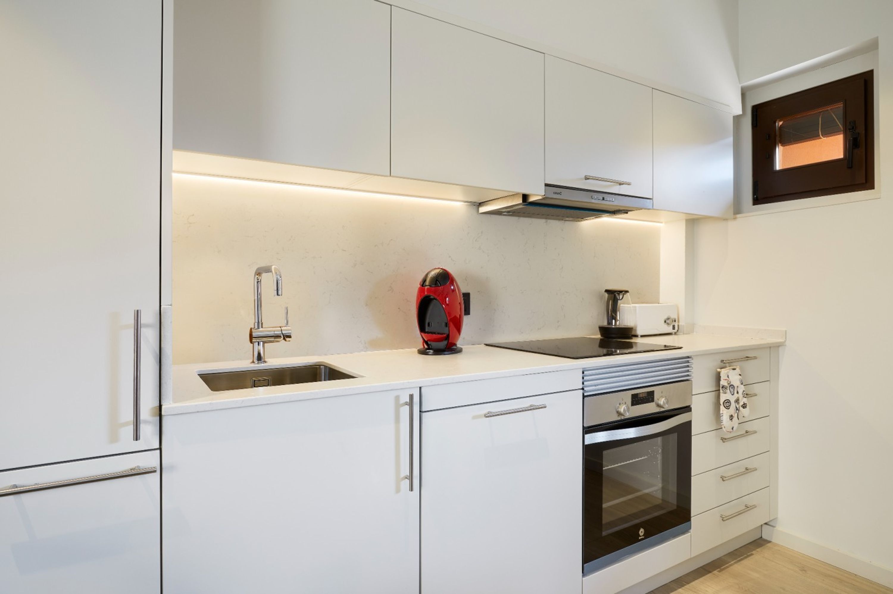 Une cuisine moderne avec des meubles blancs, un évier en acier inoxydable, une bouilloire rouge et un four.