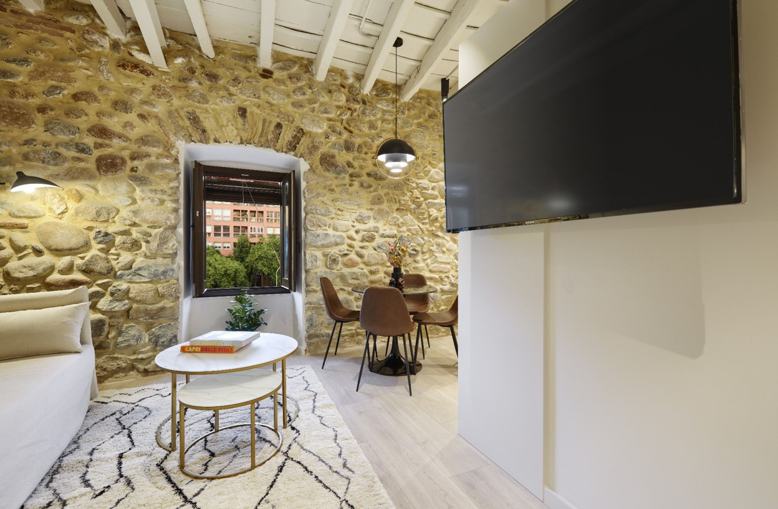A room with a stone wall, a white sofa, a round table with a book, a TV, and a dining area with chairs.
