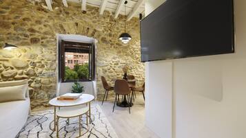 A room with a stone wall, a white sofa, a round table with a book, a TV, and a dining area with chairs.