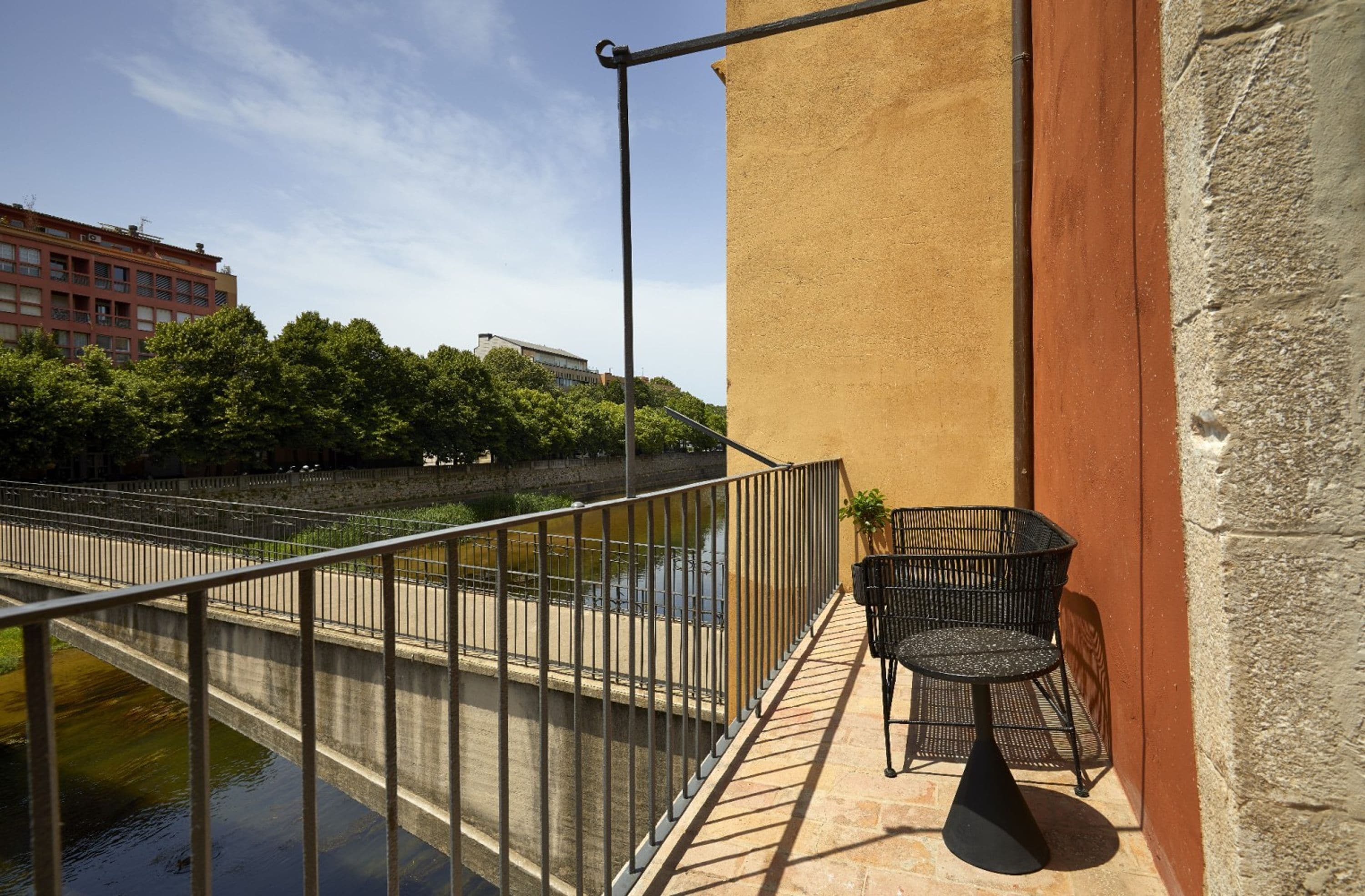 A balcony with a metal railing, a small table with chairs, and a view of a river and buildings.