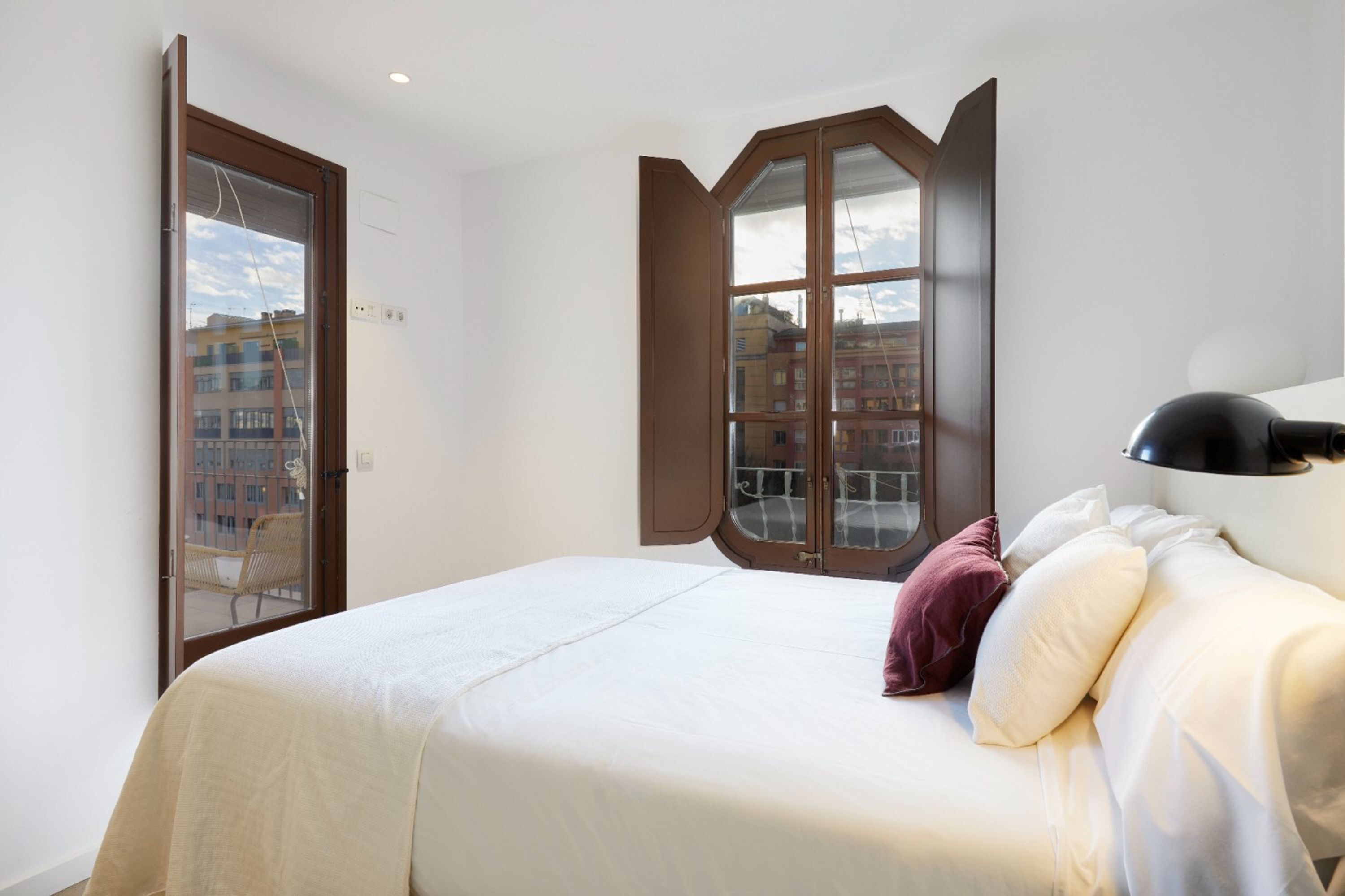 Un dormitorio con cama, almohadas, una lámpara y una ventana con vistas a edificios.