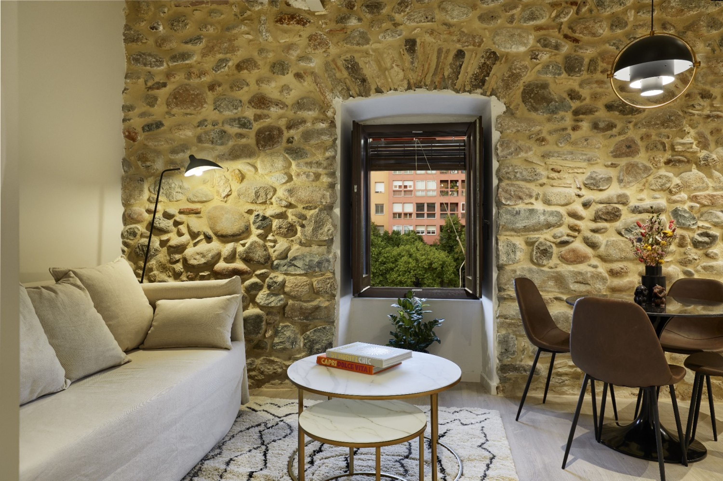 Un espacio de estar acogedor con pared de piedra, un sofá, una mesa de centro y vista a un edificio a través de una ventana.