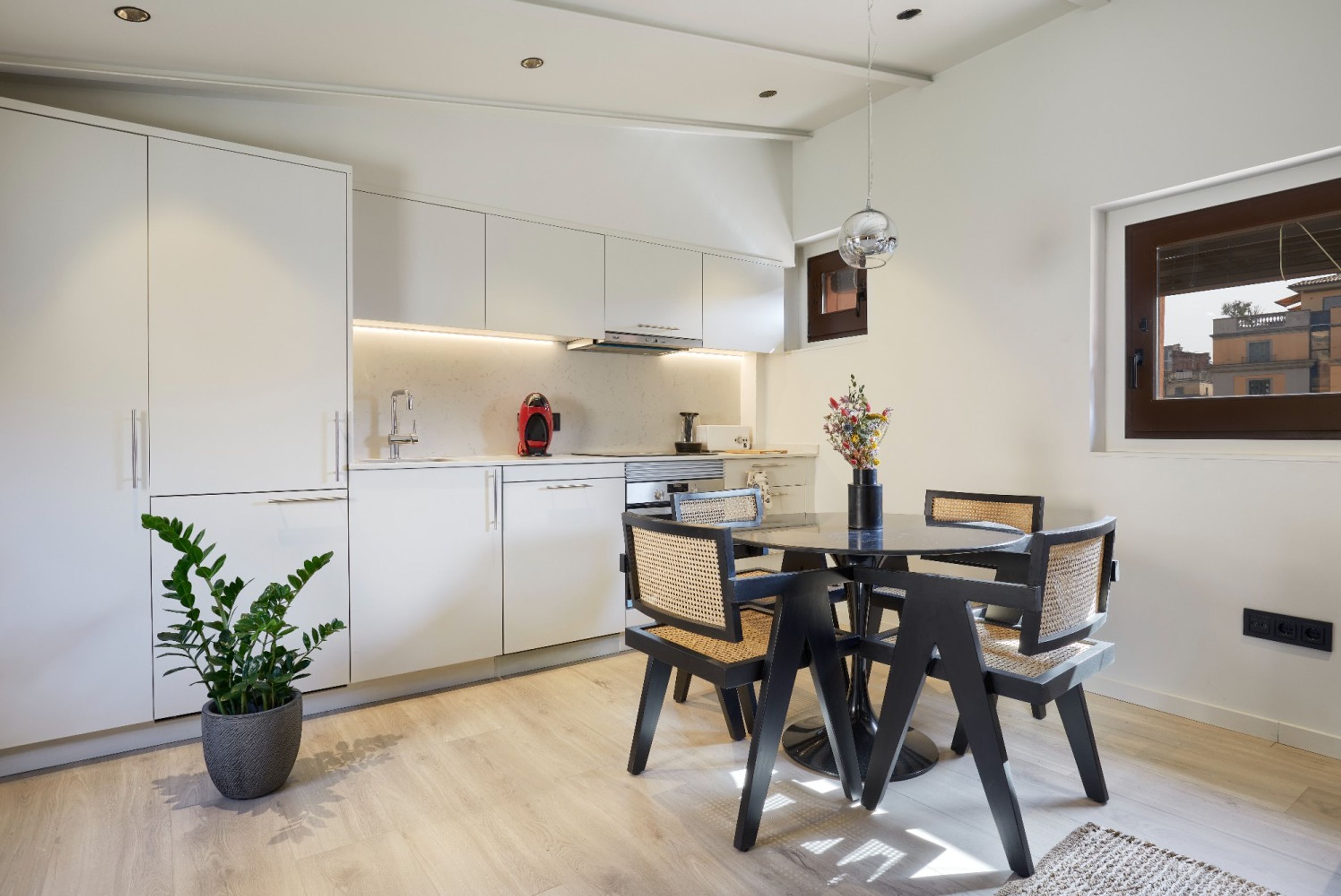 Una cocina moderna con armarios blancos, una mesa de comedor con cuatro sillas, una planta en maceta y una lámpara colgante.
