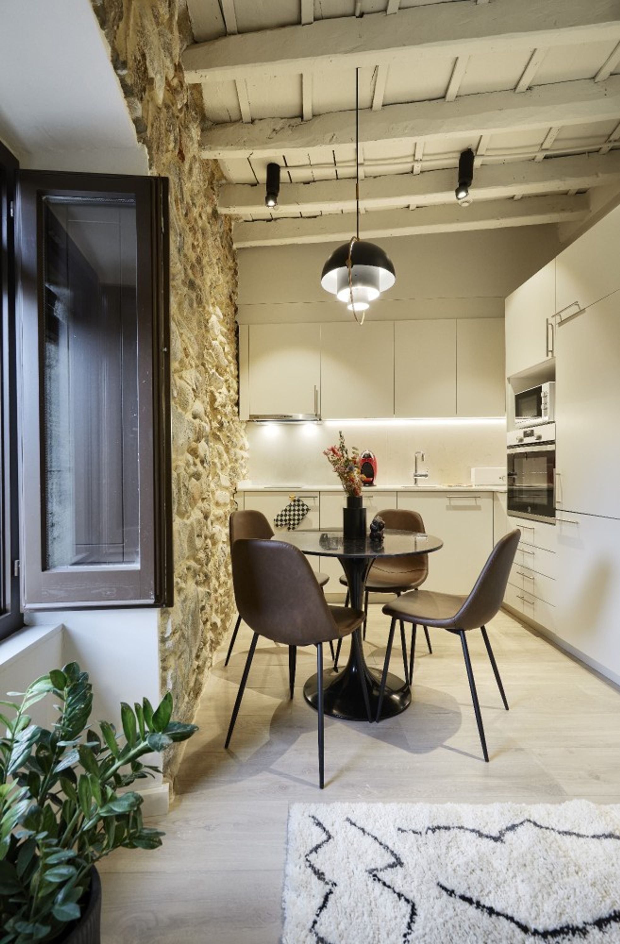 Una cocina moderna con gabinetes blancos, una mesa de comedor con sillas, una lámpara colgante y una pared decorativa de piedra.