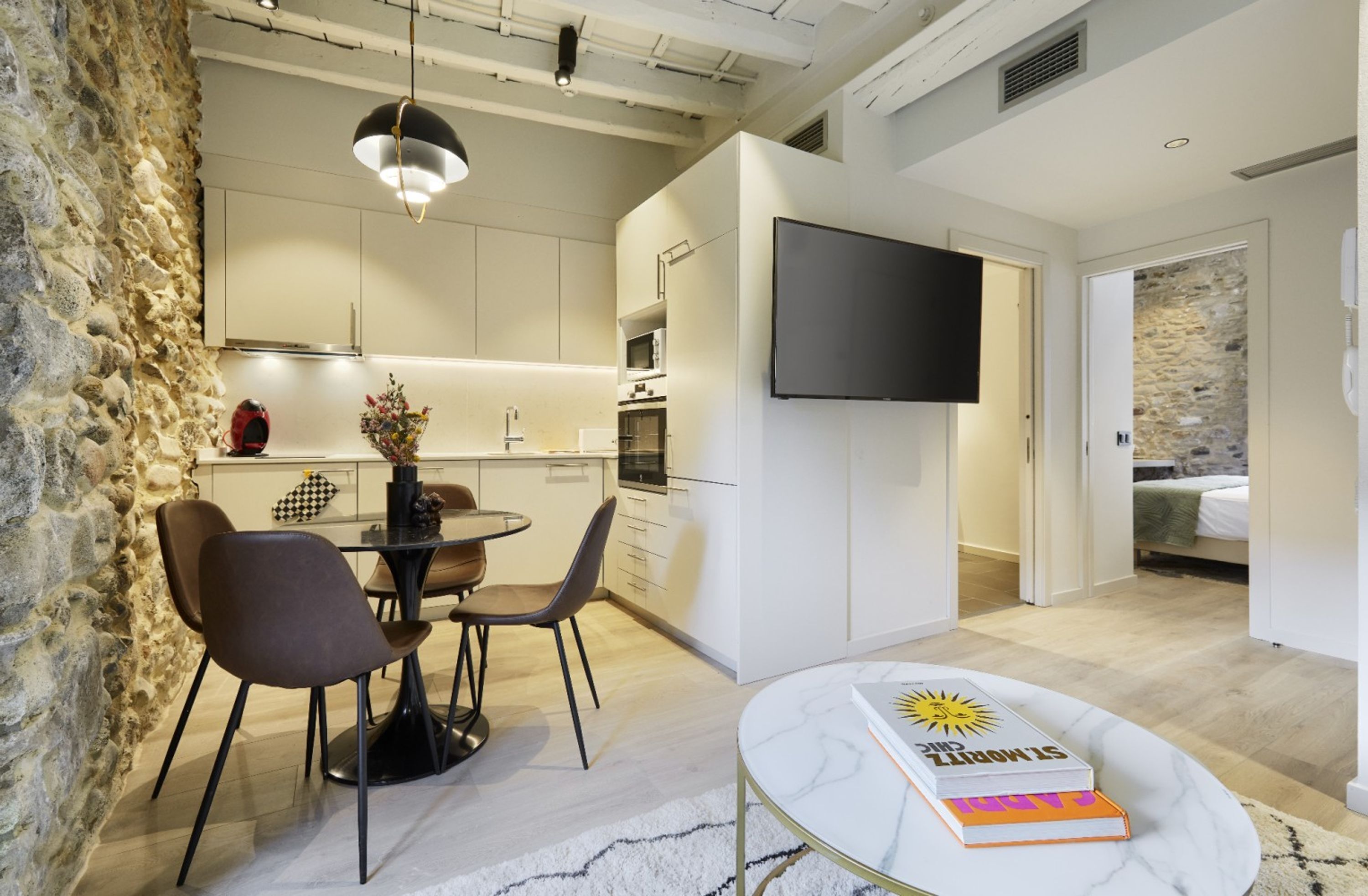 A modern kitchen with white cabinets, a dining area with a round table and chairs, a flat-screen TV mounted on the wall, and a bedroom with a bed and a stone wall.